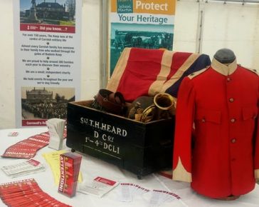 Cornwalls Regimental Museum on the Road! A display of items (including a flag, red military jacket and a bugle) arranged in a wooden chest, surrounded by leaflets, in a white marquee.