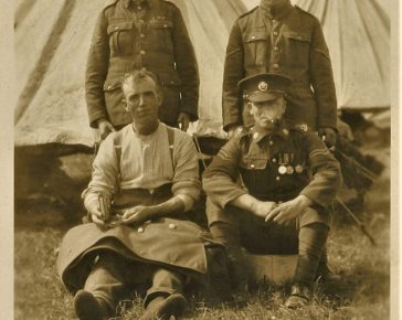 Photo of 4 non-commissioned officers – the man sitting on the left is using a sewing kit to fix the jacket which is lying across his lap